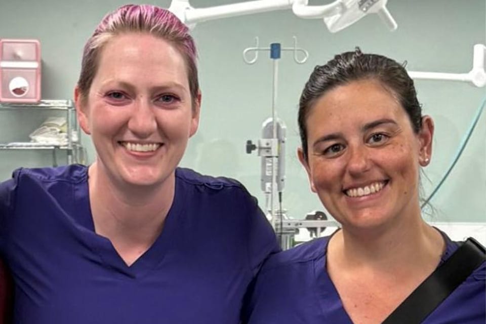 Two women wearing purple scrubs stand side by side, smiling in what appears to be a mobile vet or hospital setting, with equipment visible in the background.