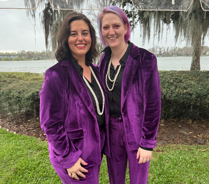 Two people smile while standing outside by a lake, both wearing matching purple velvet suits, black shirts, and pearl necklaces. Trees and greenery are in the background, giving a peaceful vibe—like finding a caring mobile veterinarian or vet near me.